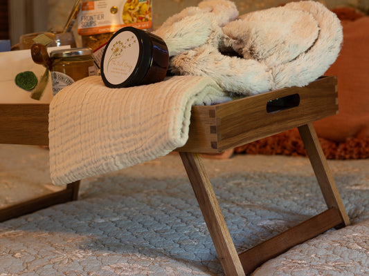Detail view of the Rest & Restore gift tray with soft Warmies slippers, a folded blanket, body butter, lemon curd, and cozy comfort items arranged on a bed.
