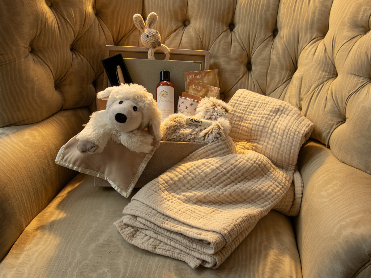 Comfort and care gift with a plush Warmies, cozy blankets, body lotion, and thoughtful treats arranged on a tufted chair.