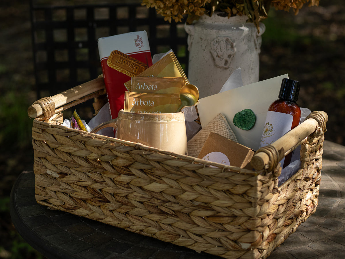 A Little Kindness gift basket with tea, biscuits, mug, soap, lotion, stationery, and small comfort items arranged outdoors on a round table.