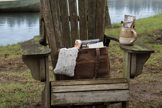 Canvas tote gift filled with Warmies neck warmer, Biscoff cookies, Bacon treat, black tea, spoon and glazed mug set on an Adirondack chair near the water