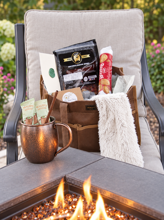 A curated outdoor fire pit gift set arranged in a brown tote bag on a patio chair beside a lit fire. The set includes Biscoff, mule mix packets in a copper mug, a Warmies neck wrap, and small gift cards, with blurred garden foliage in the background.