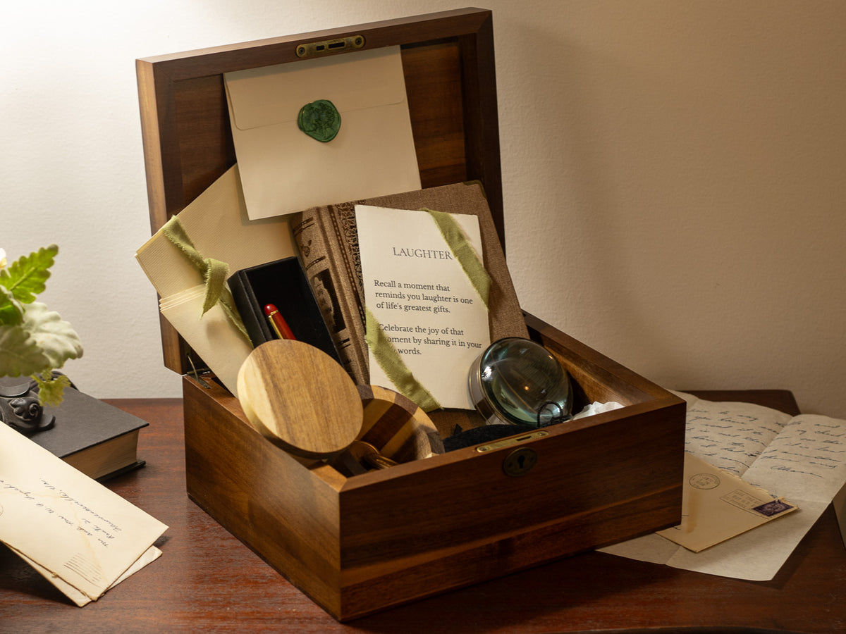 Letters & Legacy heirloom gift in an open wooden keepsake box with writing prompts, sealed stationery, pen, journal, magnifier, and vintage letters arranged on a desk.
