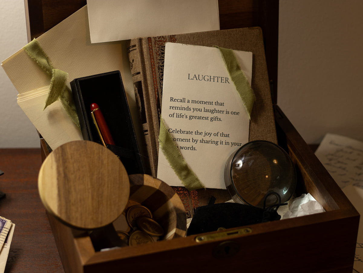 Close-up of the Letters & Legacy gift with a “Laughter” writing prompt, pen, magnifier, ribbon-tied journal, wax seal pieces, and wooden keepsake box.