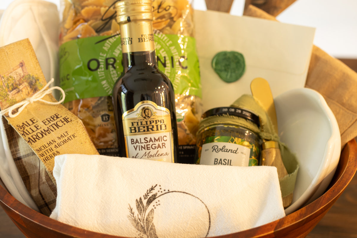 Close-up of a kitchen gift featuring balsamic vinegar, pasta, basil, aromatic salt, serving pieces, and a folded kitchen towel arranged in a wooden bowl.