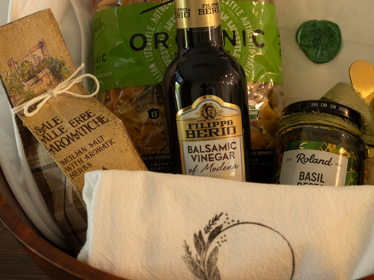 Close-up of the kitchen gift with balsamic vinegar, basil, aromatic salt, pasta, serving bowl, kitchen linen, and wooden bowl.