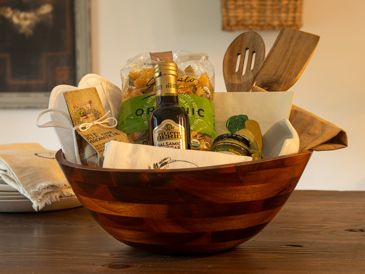 Kitchen gift arranged in a large wooden bowl with pasta, balsamic glaze, wooden utensils, kitchen linens, seasoning, and serving pieces.