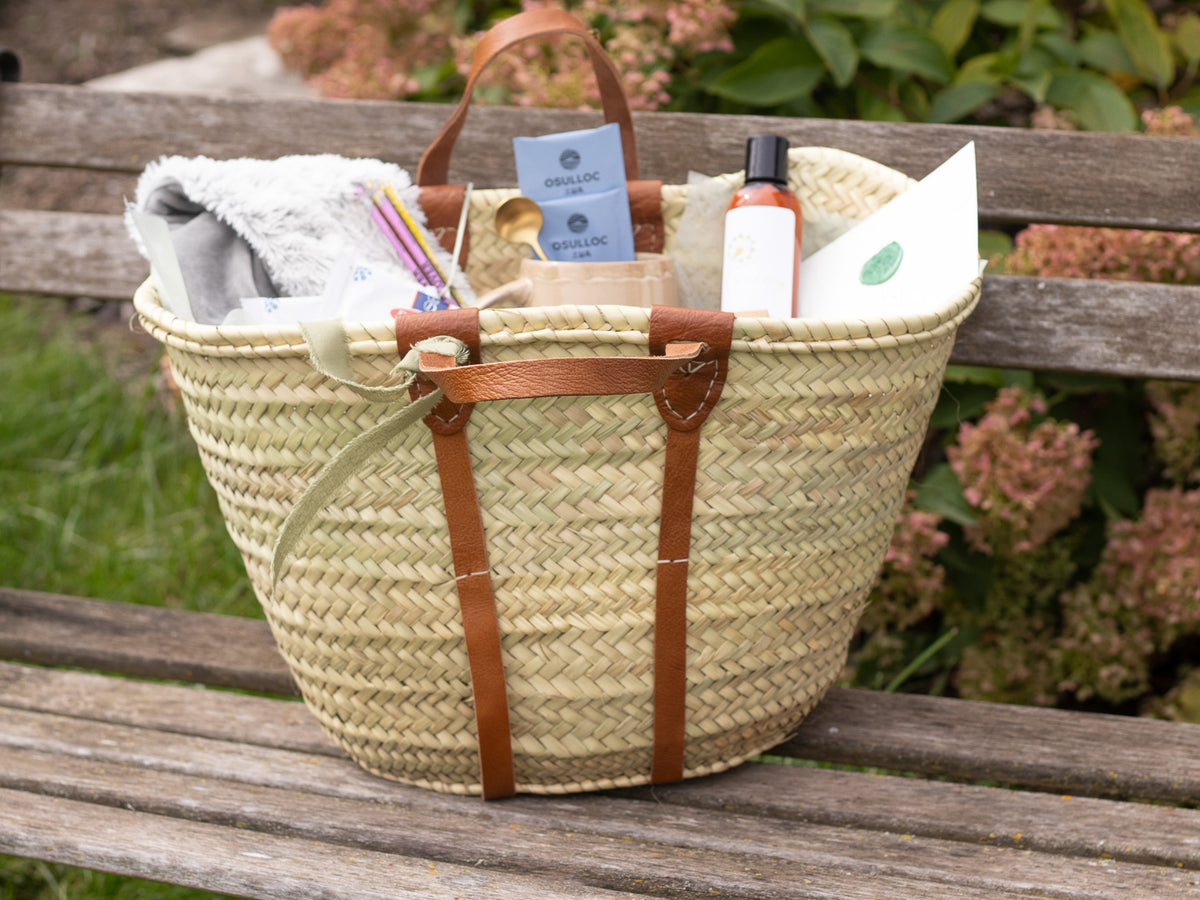 Irish Daisy Quiet Moment Gift in woven straw tote gift basket sits on a weathered garden bench, filled with soft spa comforts, stationery, a pen, lotion, and thoughtful Irish Daisy details in soft sage and cream ton