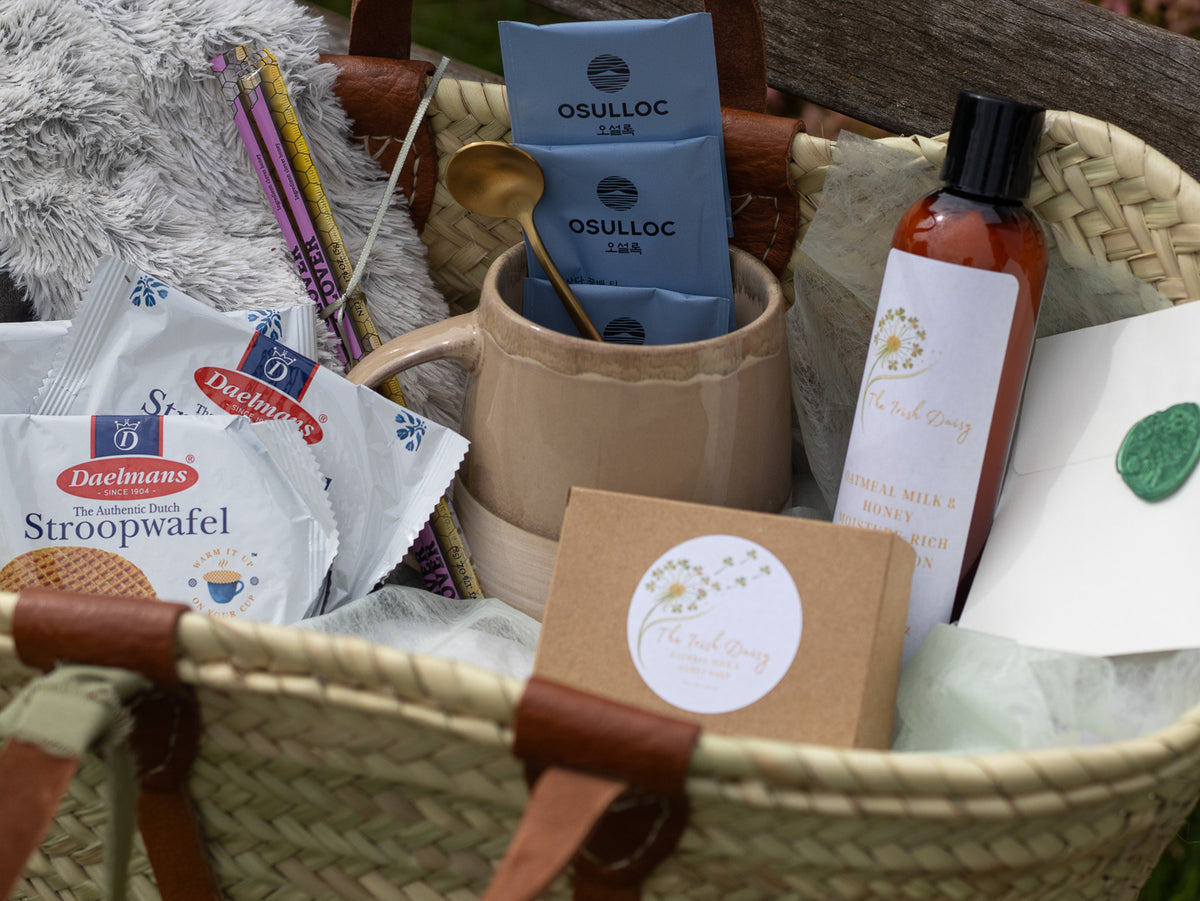 Close-up of a woven gift tote with tea, stroopwafels, mug, body lotion, stationery, and a sealed note arranged inside.