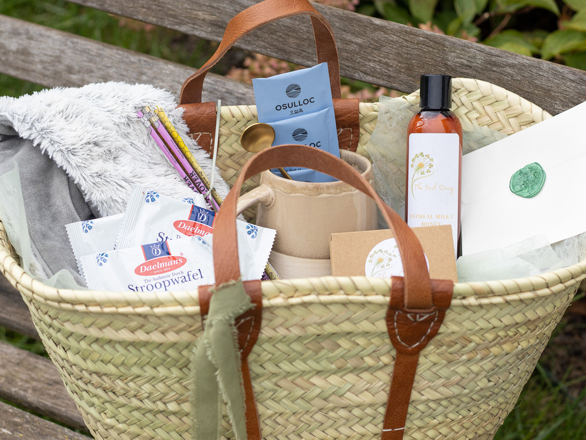 Close-up of a woven gift tote with body butter, lotion, stationery, a pen, and soft comfort items arranged inside.