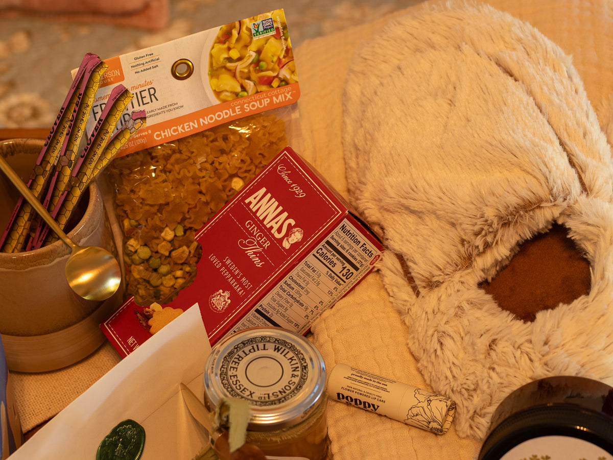 Close-up of the Rest & Restore gift with chicken noodle soup, ginger cookies, honey sticks, mug, soft plush wrap, and comfort-care items.