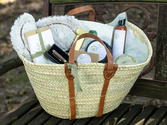 Woven gift tote with leather handles filled with stationery, a pen, body care items, soft comfort pieces, and thoughtful keepsakes displayed on a garden bench.