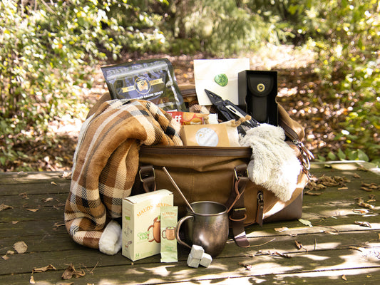 Heritage Outdoorsman Weekender gift with a waxed canvas bag, plaid blanket, metal mug, and gourmet treats in a wooded setting.