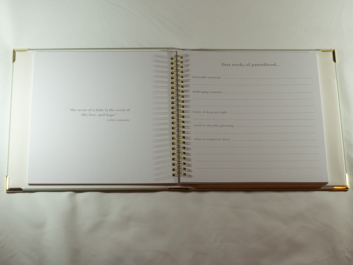 Open baby memory book showing guided prompts for the first weeks of parenthood, with gold spiral binding and corner details.
