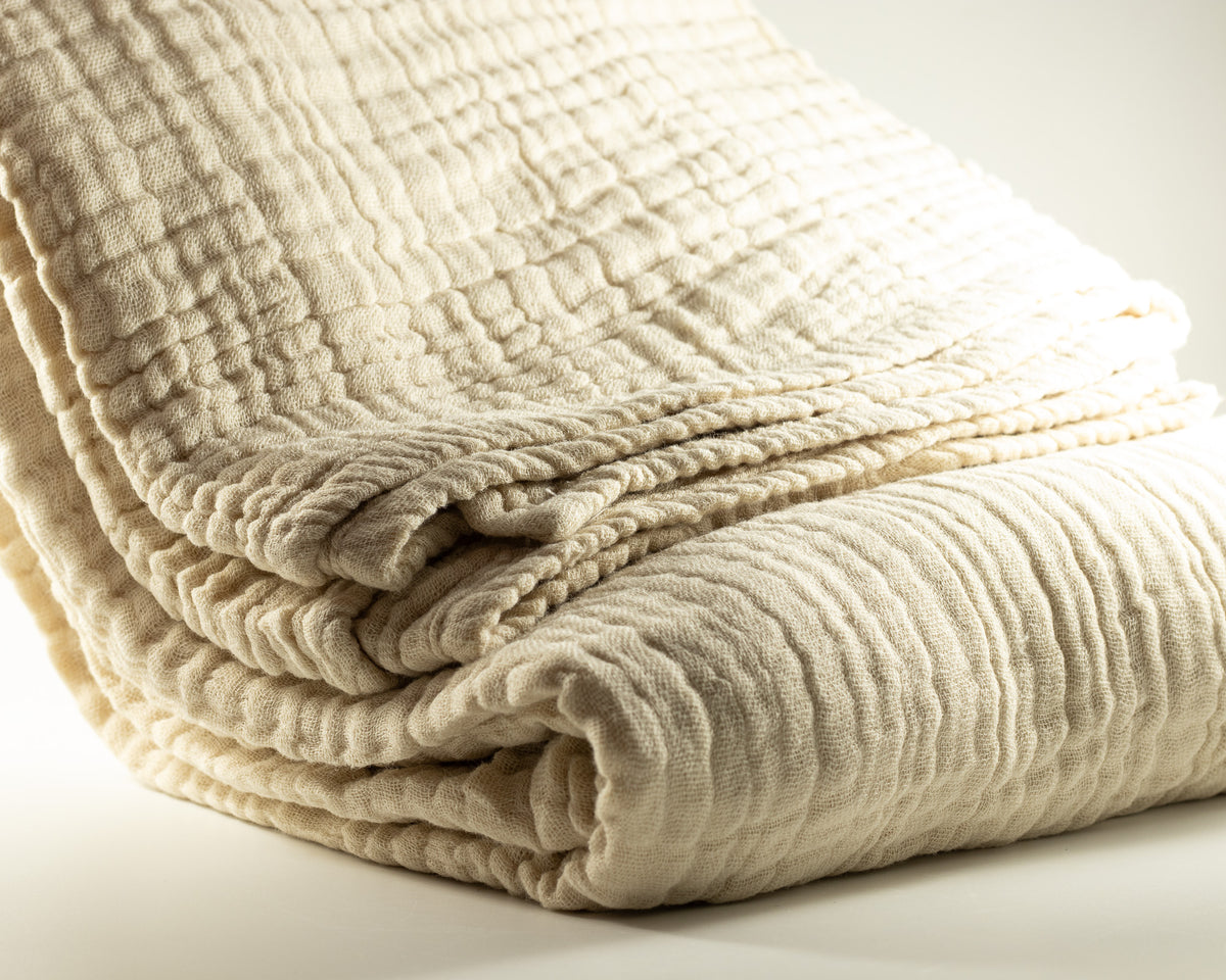 A folded cream-colored Irish Daisy muslin blanket with a textured, crinkled gauze pattern, photographed on a clean white background for gift basket.