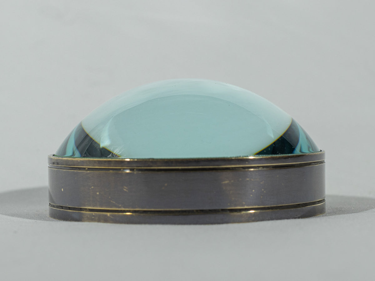 Vintage blue-green glass magnifying paperweight with an aged brass-tone base, photographed on a soft neutral background.