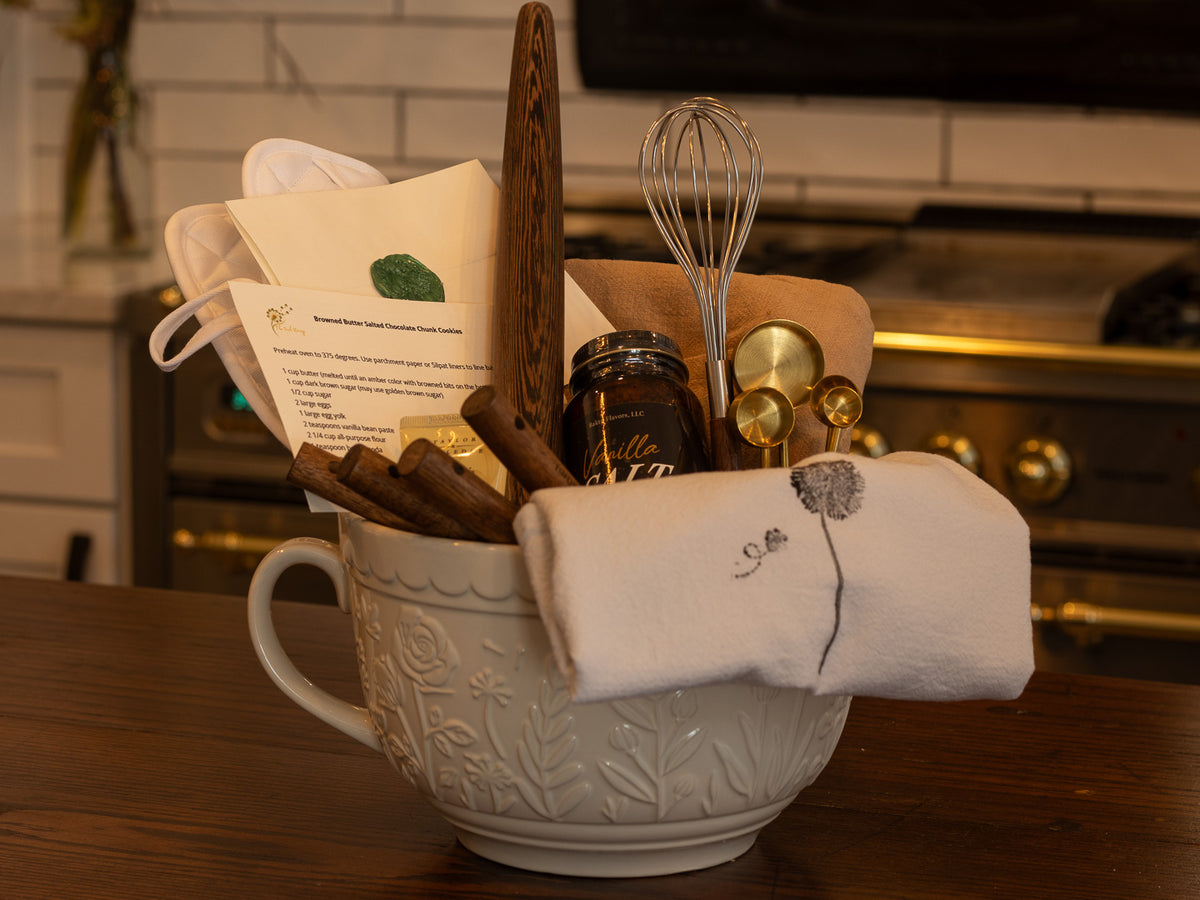 Kitchen gift arranged in a ceramic mixing bowl with linens, utensils, recipe card, measuring spoons, whisk, and culinary treats on a wooden table.