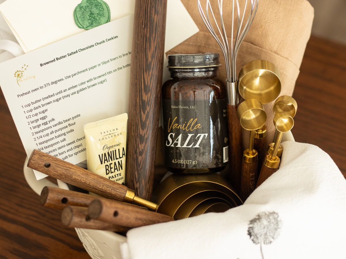Kitchen gift arranged in a ceramic mixing bowl with linens, utensils, recipe card, measuring spoons, whisk, and culinary treats on a wooden table.